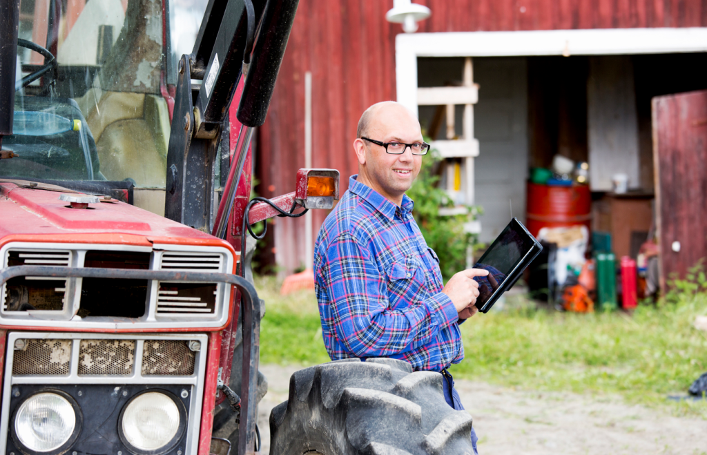 Bilden visar en lantbrukare som står vid sin traktor och håller i en läsplatta. 